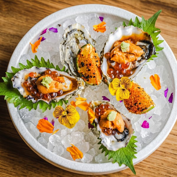Four oysters with uni, roe, and wasabi, garnished with edible flowers on a bed of ice.