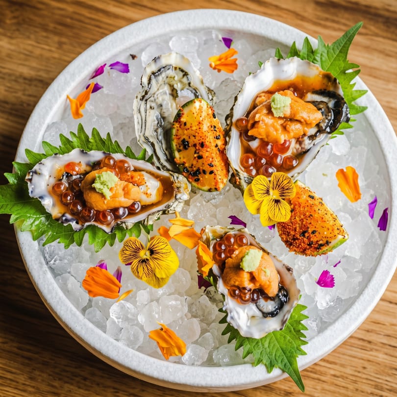 Four oysters with uni, roe, and wasabi, garnished with edible flowers on a bed of ice.