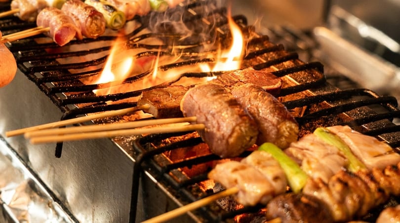 Skewers of meat and vegetables grilling over an open flame with smoke rising.