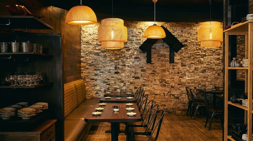 Cozy restaurant interior with bamboo pendant lights, brick wall, wooden tables, and banquette seating.
