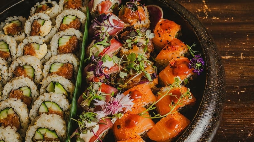 A platter of various sushi rolls, including cucumber and spicy tuna, on a dark wooden surface.