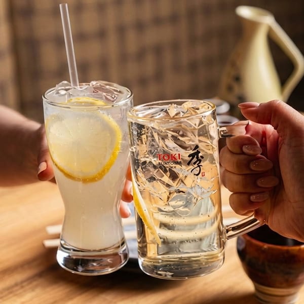 Two hands clinking glasses of refreshing drinks with lemon slices and ice on a wooden table.