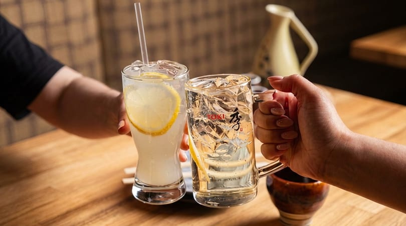 Two hands clinking glasses of refreshing drinks with lemon slices and ice on a wooden table.