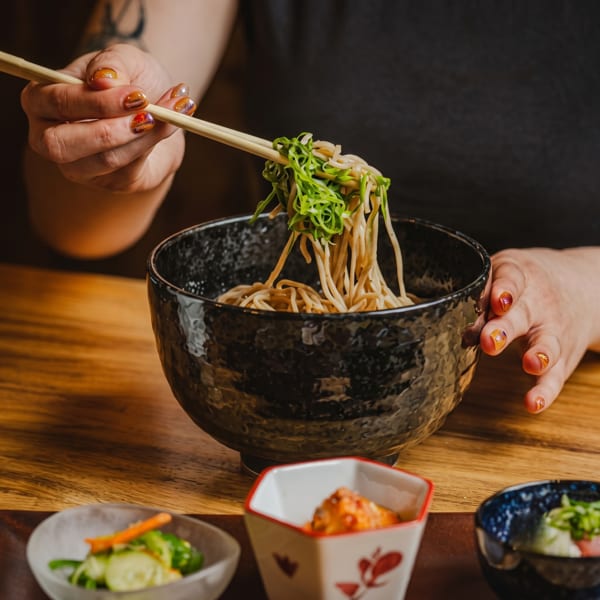 Hands with painted nails using chopsticks to lift noodles and green onions from a black bowl.